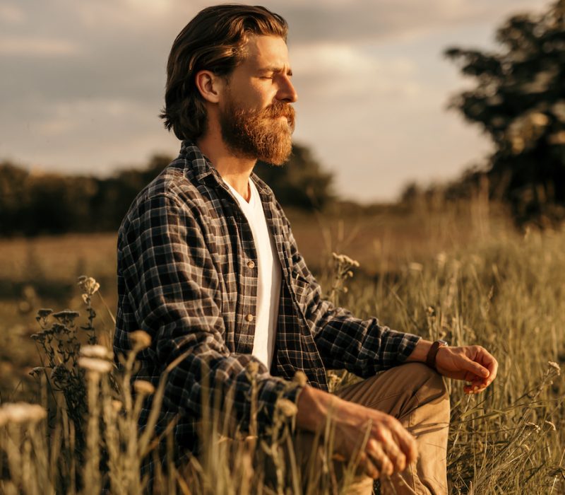 Relaxed bearded man with closed eyes sitting in meadow and meditating while establishing connection with nature in cloudy evening